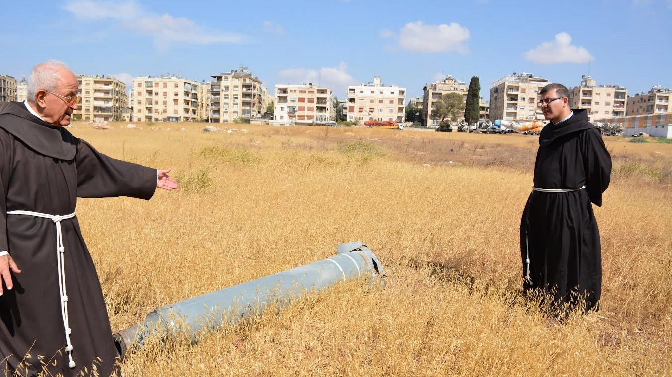 Padre Firas Lutfi (a destra) davanti a un razzo. Sullo sfondo la città di Aleppo