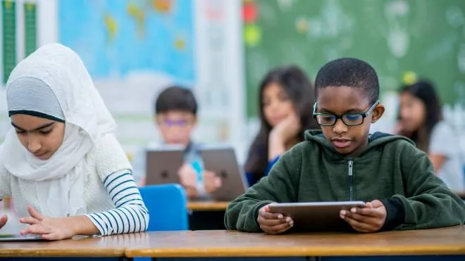 A Muslim girl and a boy of African descent are sitting beside each other in a classroom. They are working with tablet computer. They are learning about technology.